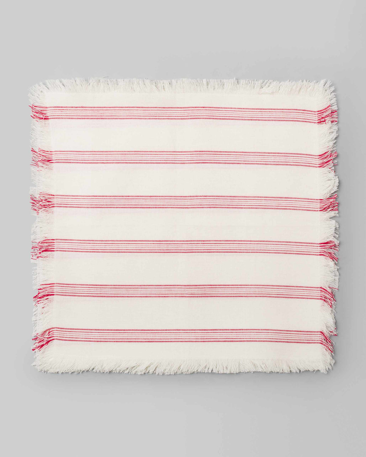Shima Dinner Napkin in ivory with red stripes and fringed edges, laid flat.