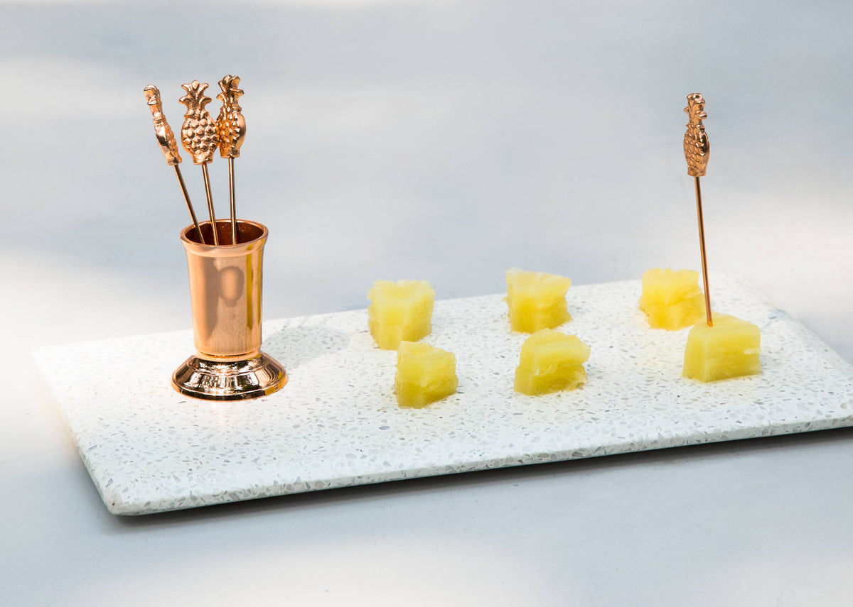 Anasi fruit picks in a copper stand, beside a tray of pineapple pieces.