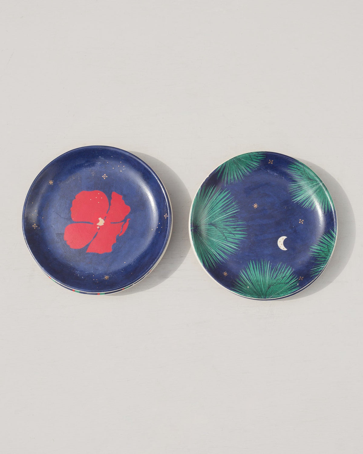 Two Wonderland Tea Plates, one with a red hibiscus and one with palm leaves and a moon.