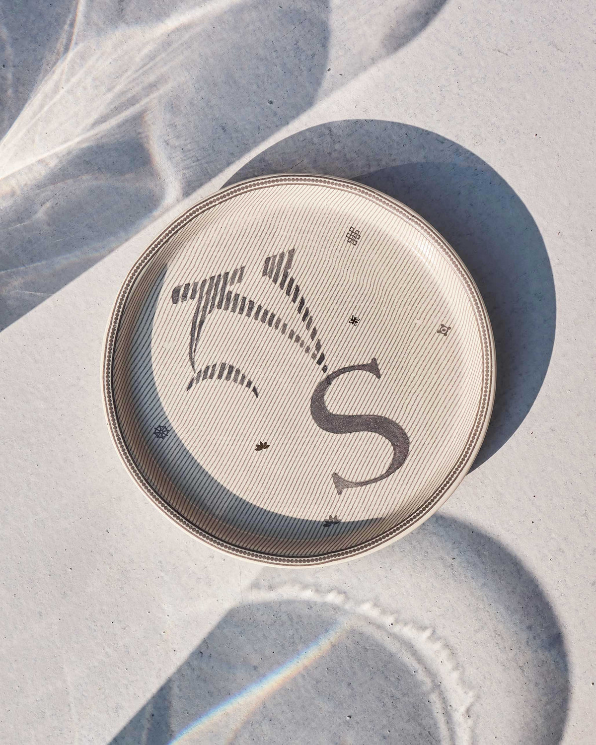 Cream Akshar coaster with a charcoal letter S and small lotus accents.