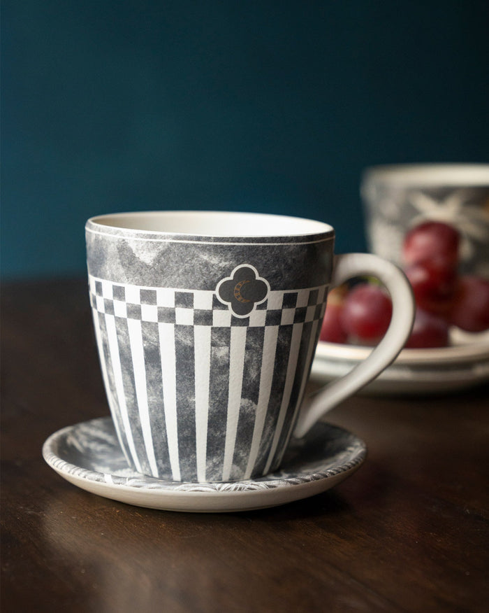 Mhadei mug in grey and white with a checked band, beside a saucer.