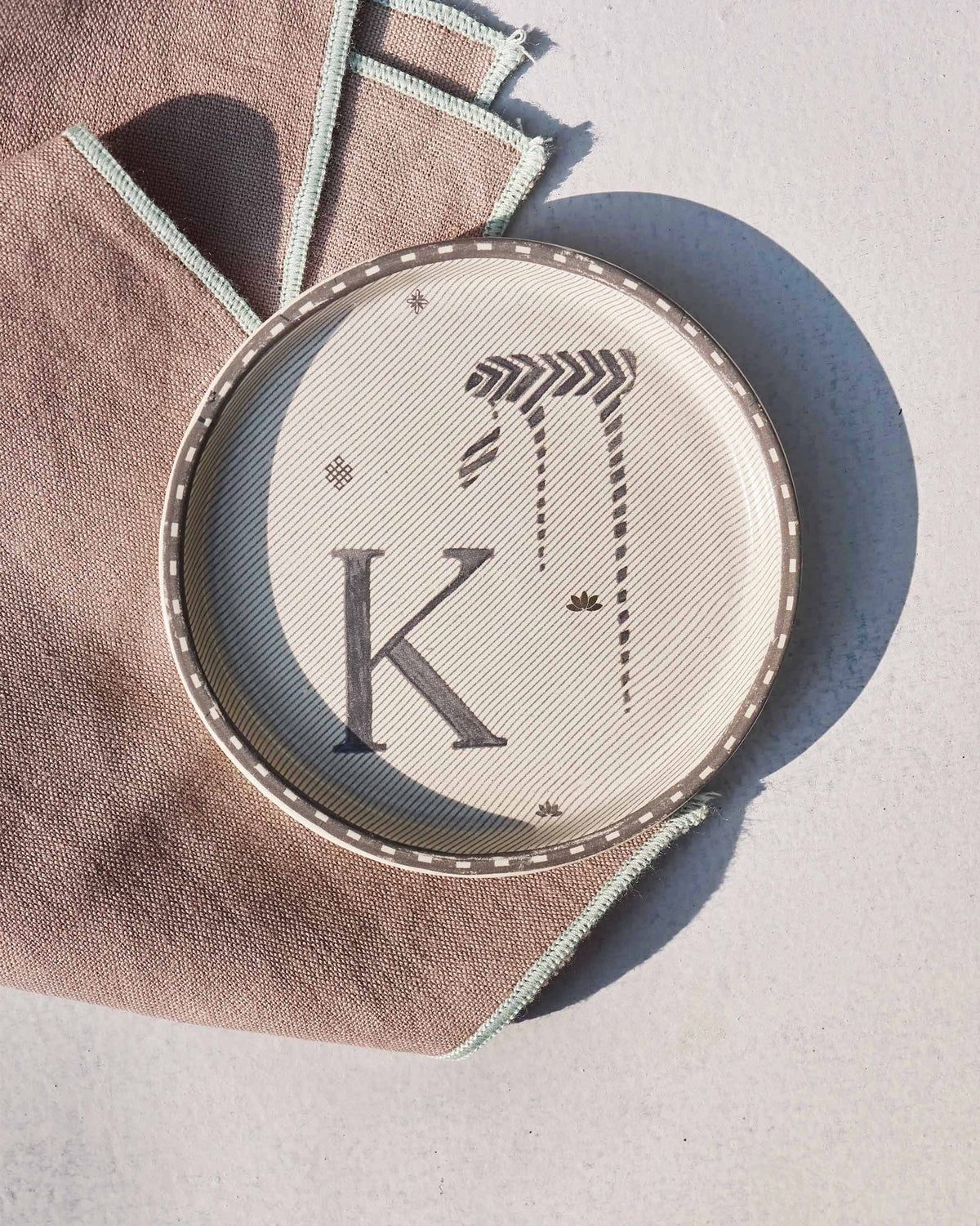 Akshar Coaster with a letter K and a striped glyph on cream.