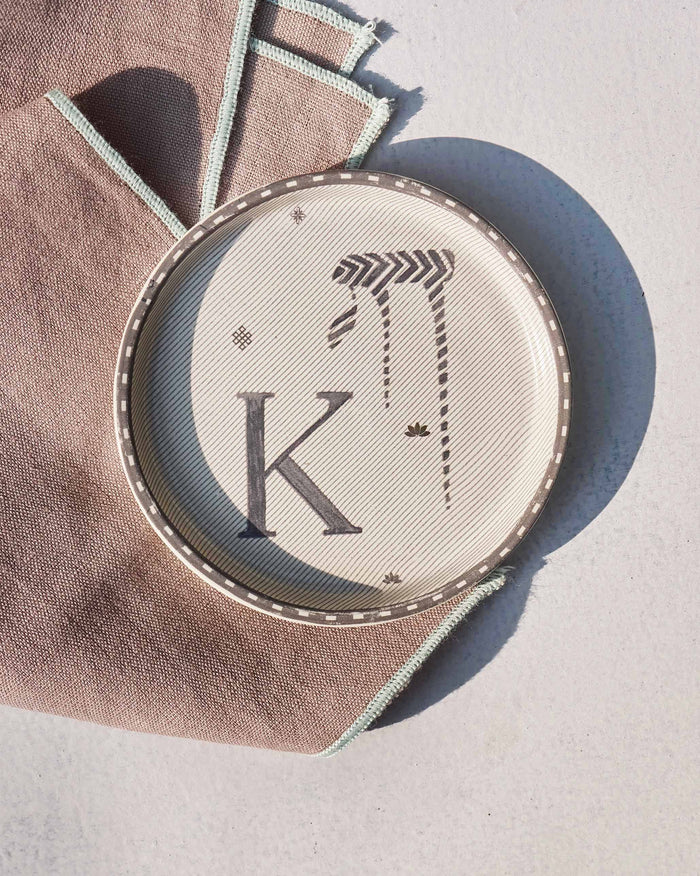 Akshar Coaster with a letter K and a striped glyph on cream.
