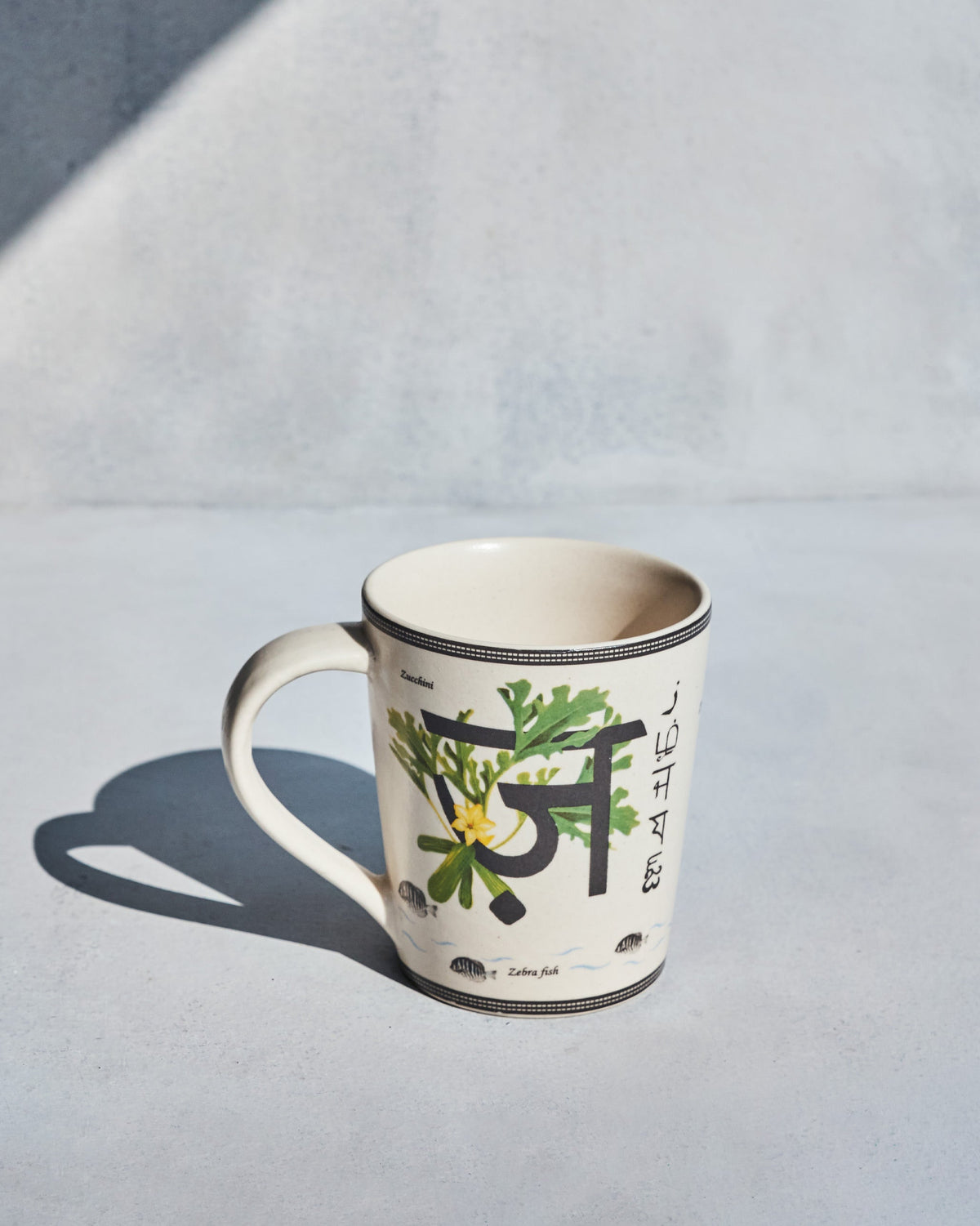 Cream Zafran mug with a Devanagari letter, zucchini leaves and zebra fish motifs.