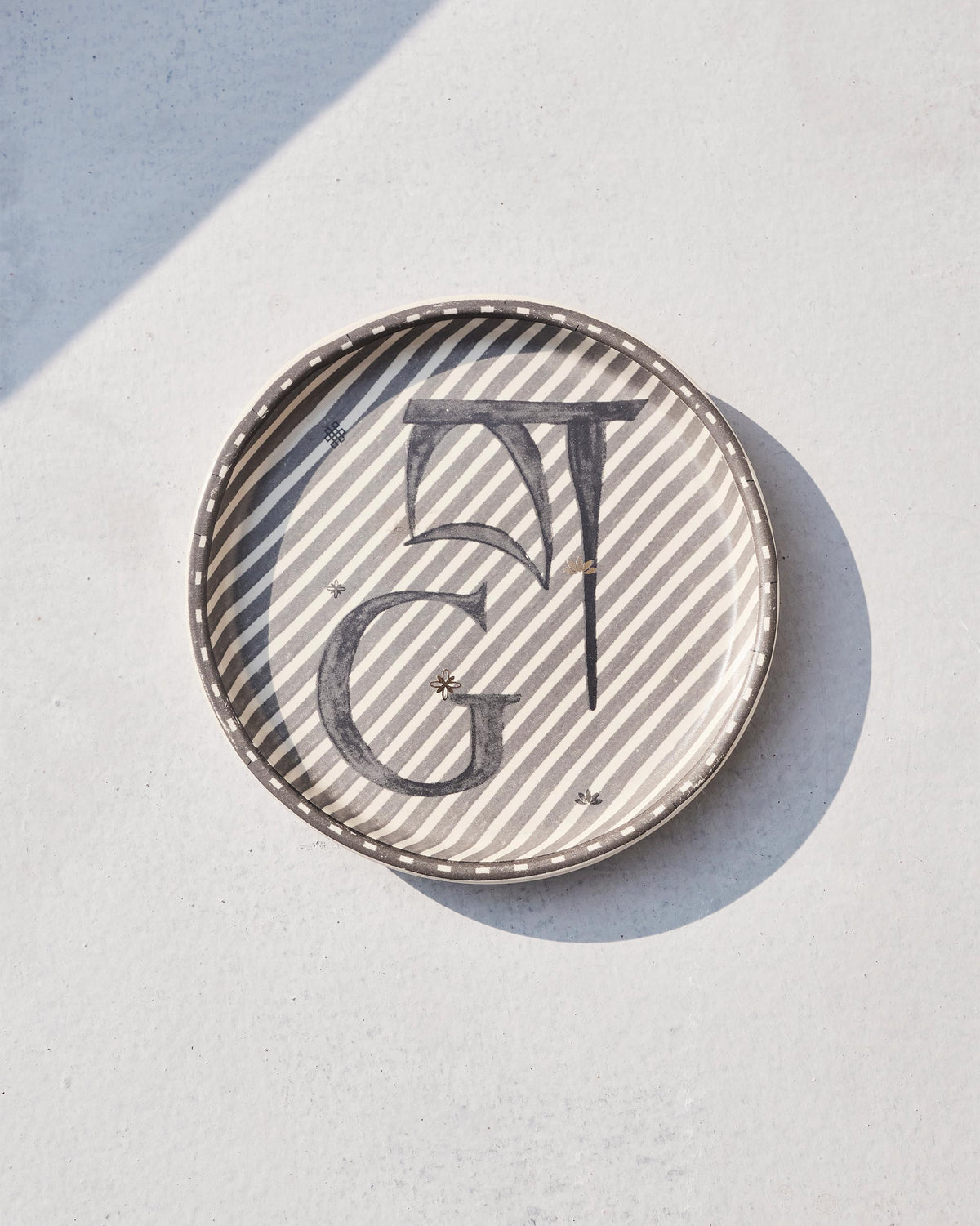 Akshar Coaster with a letter G on grey diagonal stripes, seen at an angle.