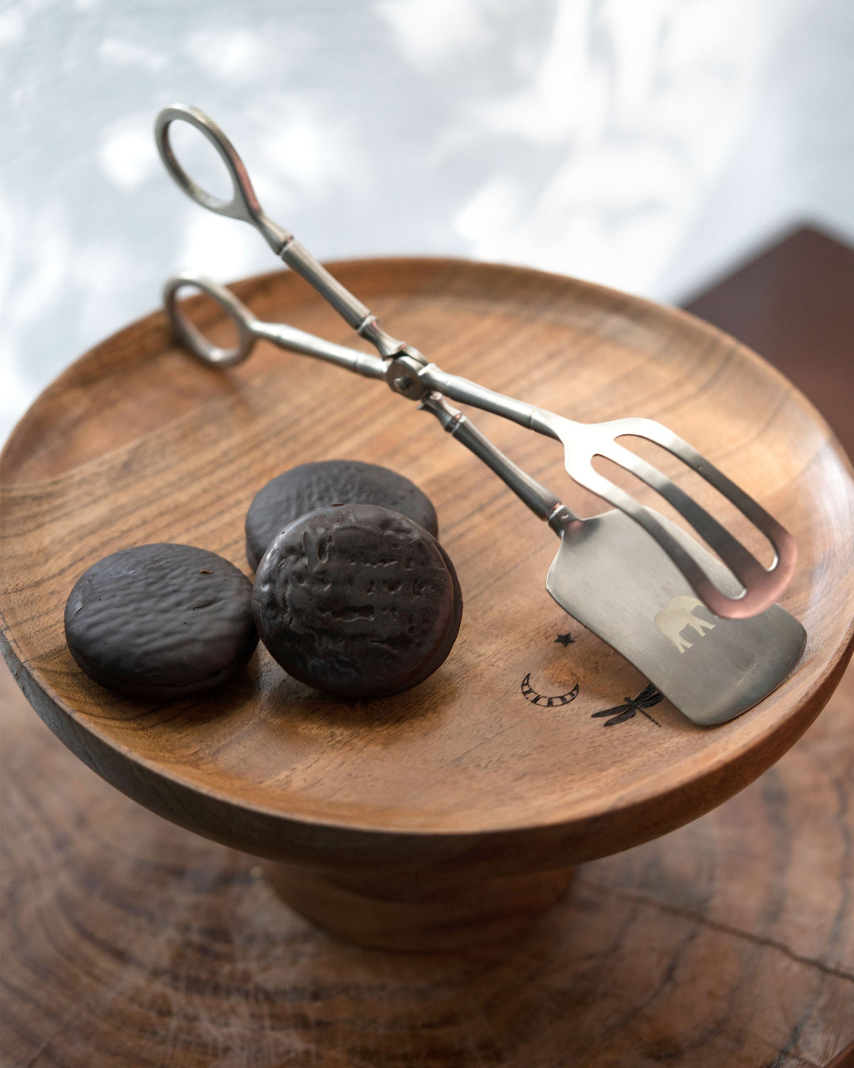 Celeste Dessert Server tongs resting on a wooden stand beside dark biscuits.
