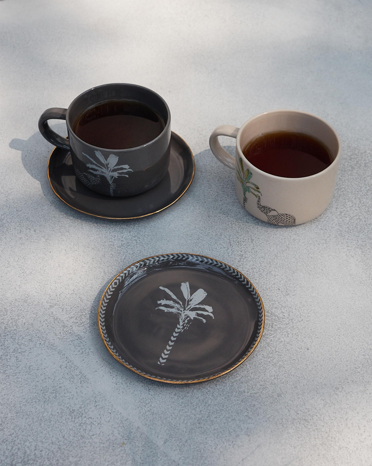 Coastal Tea Plates in charcoal with a palm motif, styled beside tea cups and a saucer.