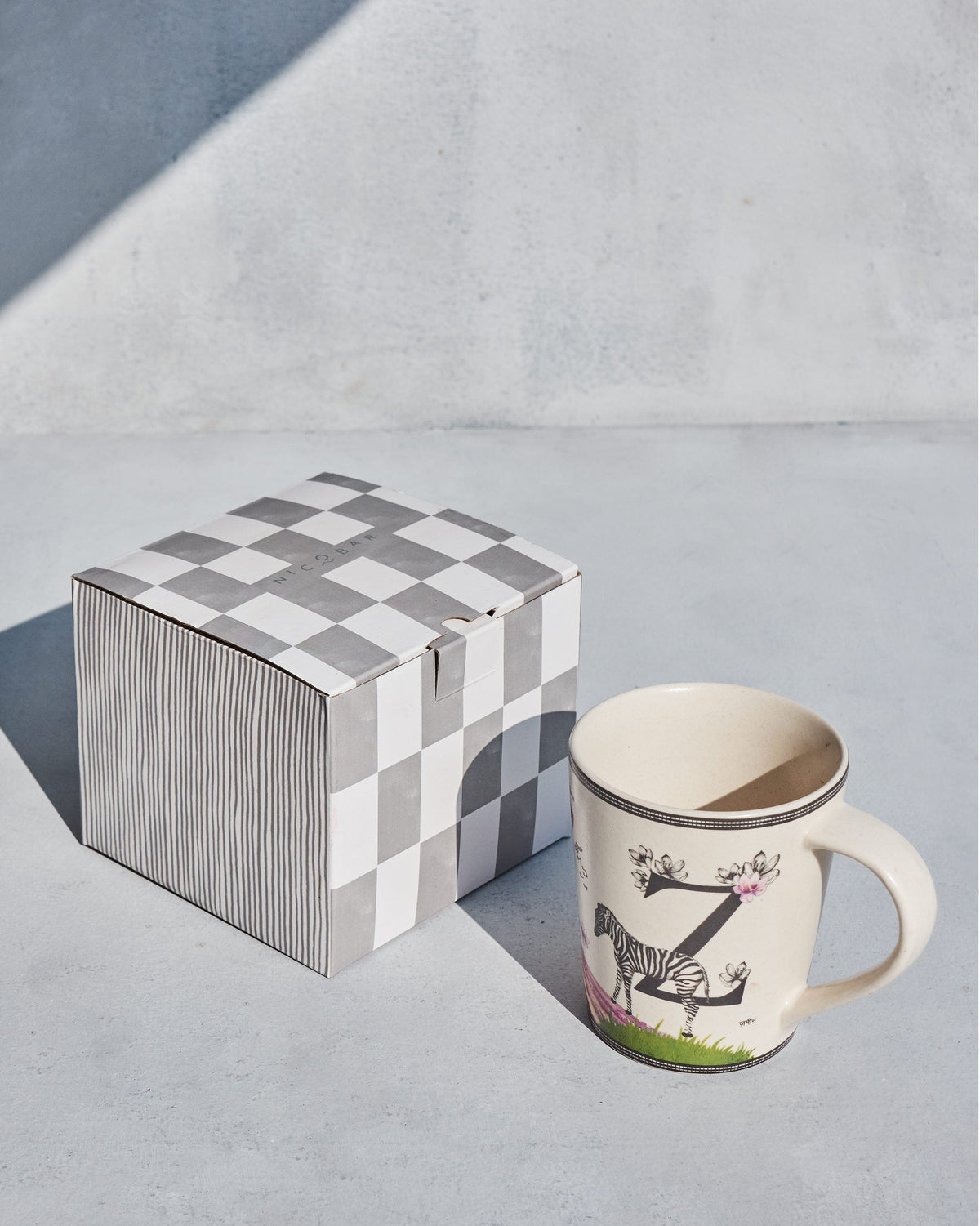 Cream Zafran alphabet mug beside a grey-and-white checked Nicobar gift box.
