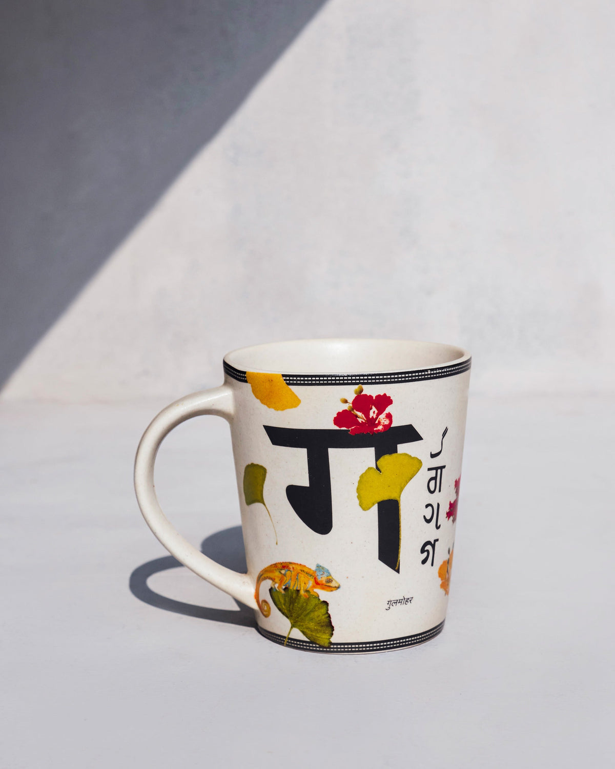 Gulmohar mug reverse with Devanagari ग, a red flower, and a small chameleon.