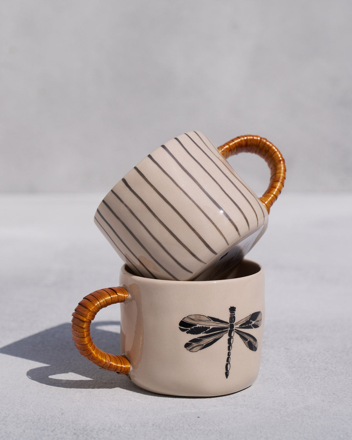 Cream dragonfly mug stacked with a striped mug, both with cane-wrapped handles.