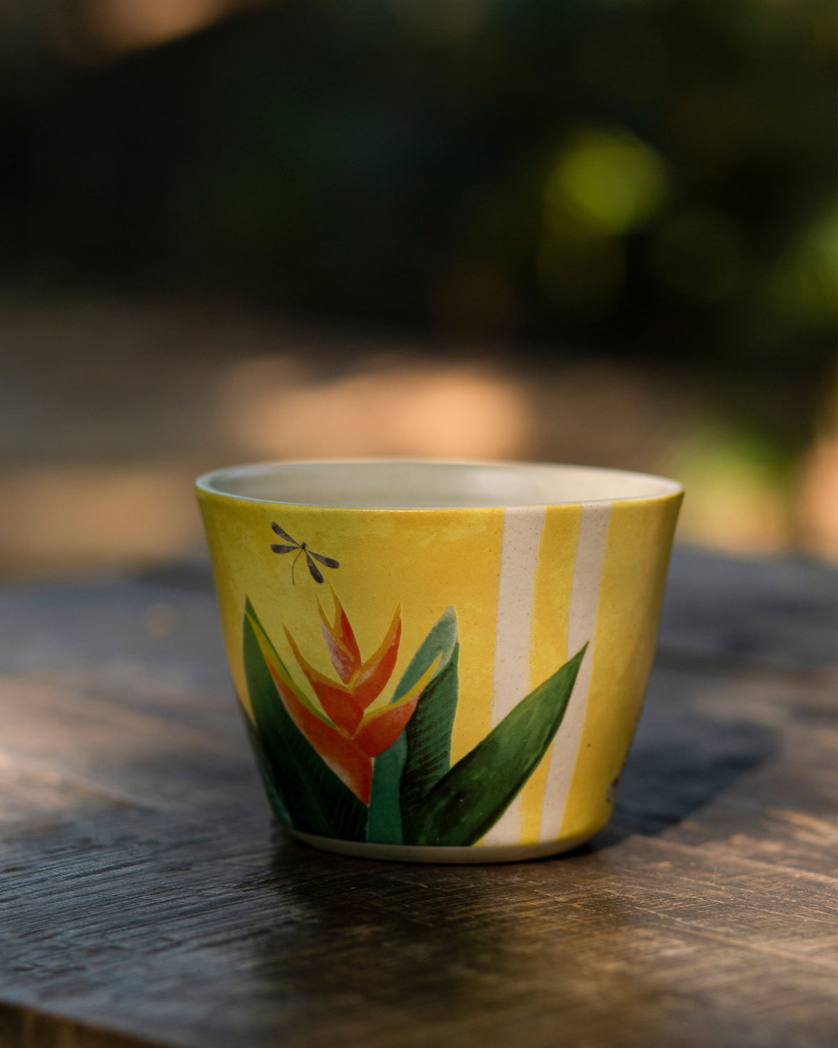 Yellow cup with a heliconia flower and dragonfly print on a wooden table.