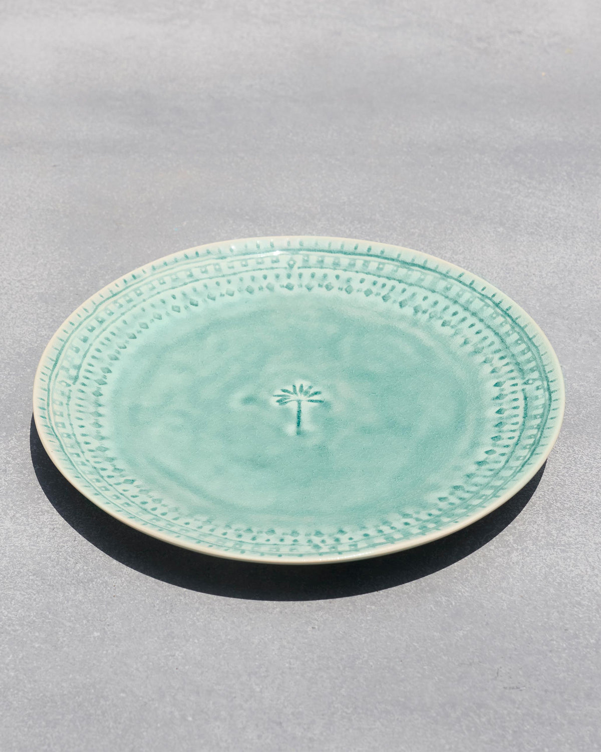 Coco Palm Dinner Plate in aqua, viewed at an angle on a grey surface.
