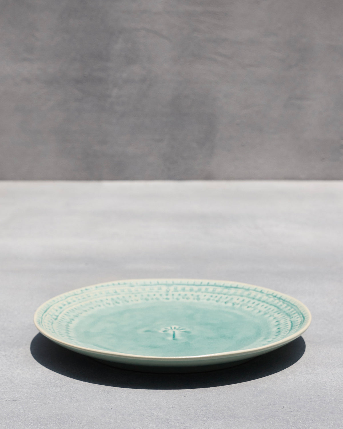 Side view of Coco Palm Dinner Plate in aqua on a grey surface.