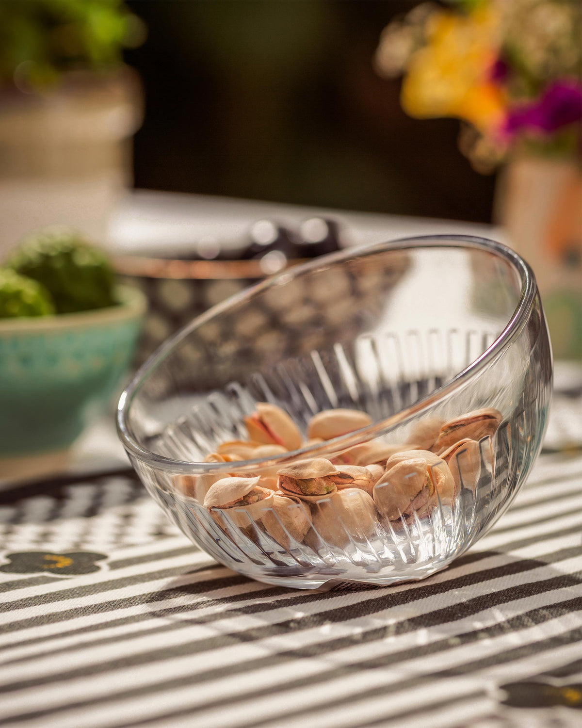 Clear fluted Mahi nut bowl holding pistachios on a striped cloth.
