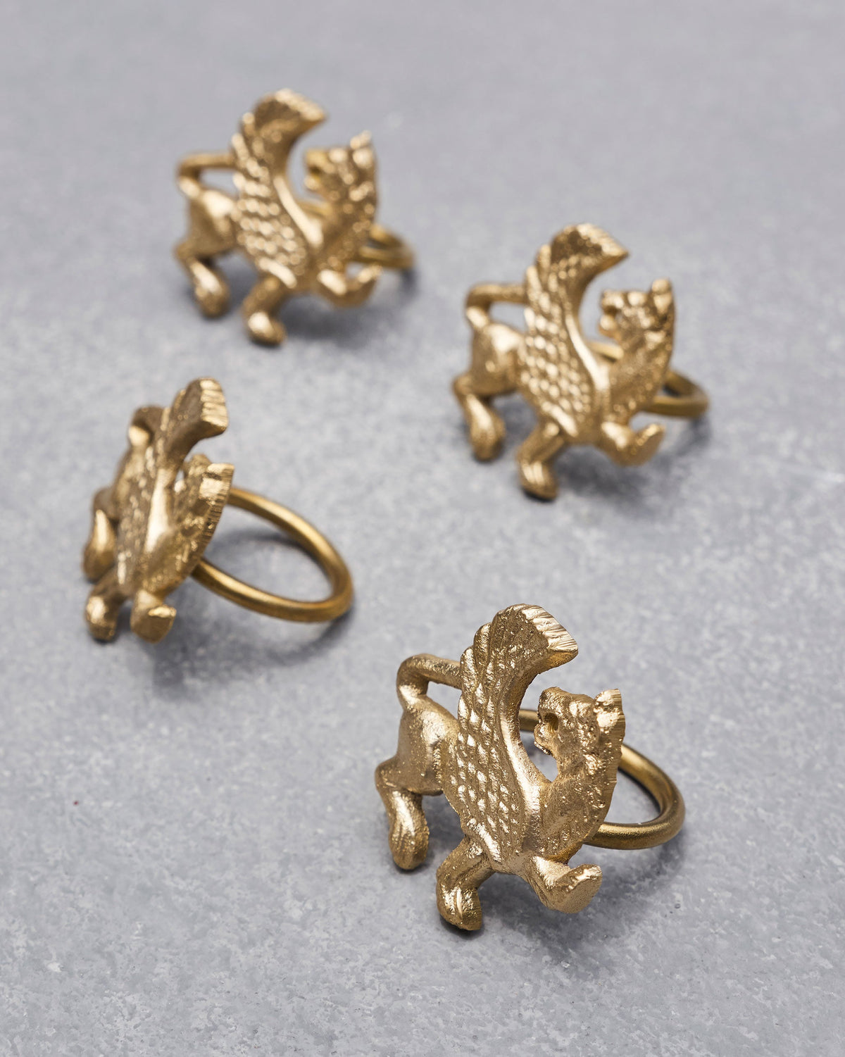 Four brass Chinthe Napkin Rings scattered at angles on a grey surface.