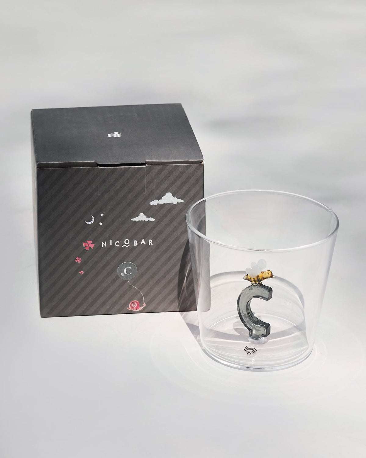 Clear Alphabet glass with a 3D letter C beside a charcoal Nicobar gift box.