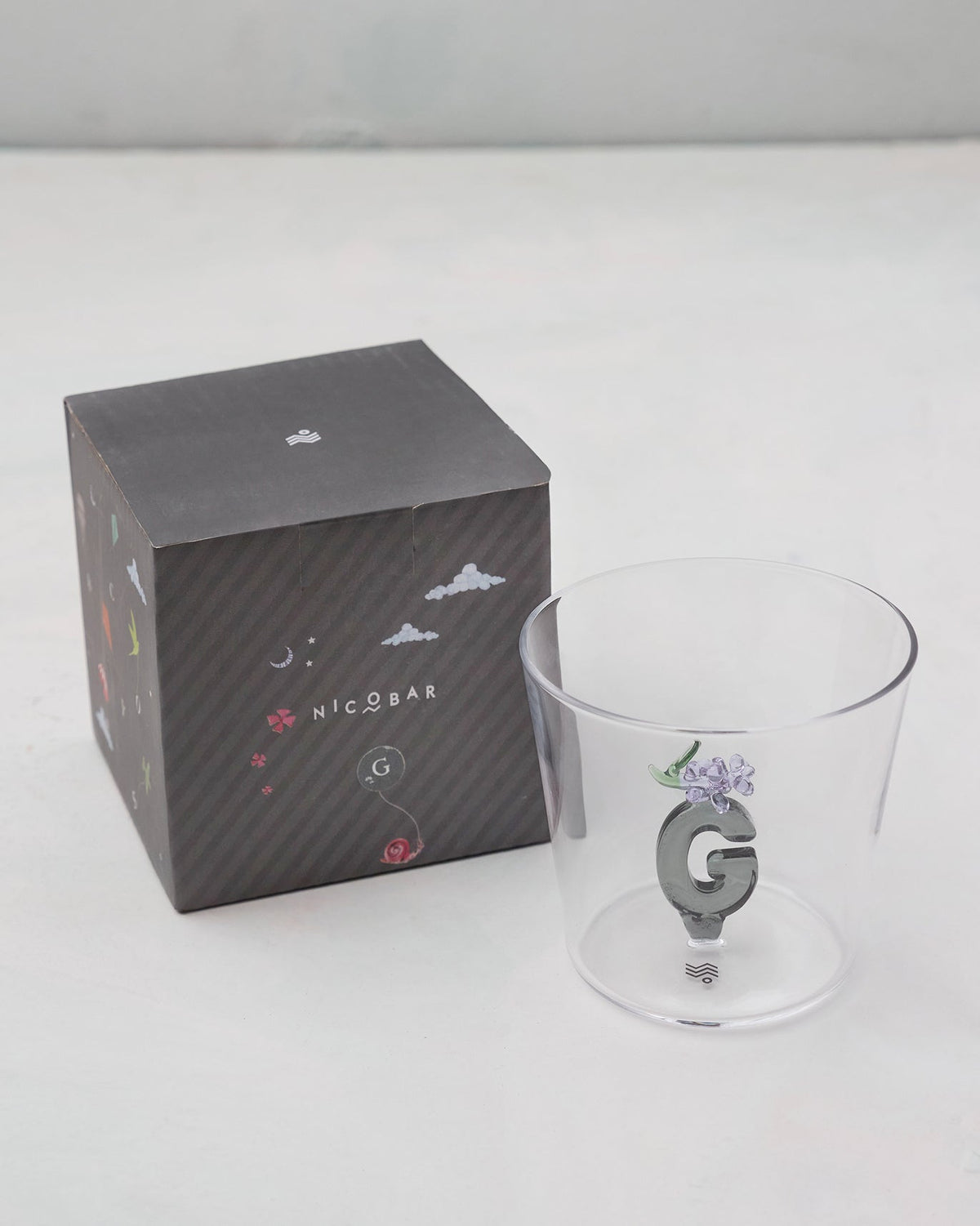 Clear Alphabet glass with a 3D letter G beside a charcoal Nicobar gift box.