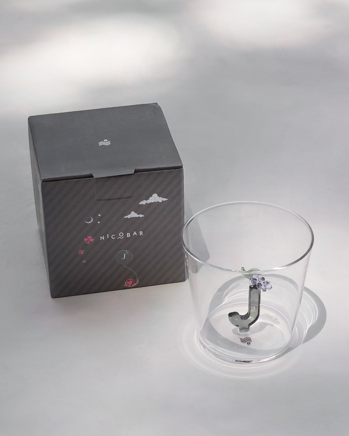 Clear Alphabet glass with a 3D letter J beside a charcoal Nicobar gift box.