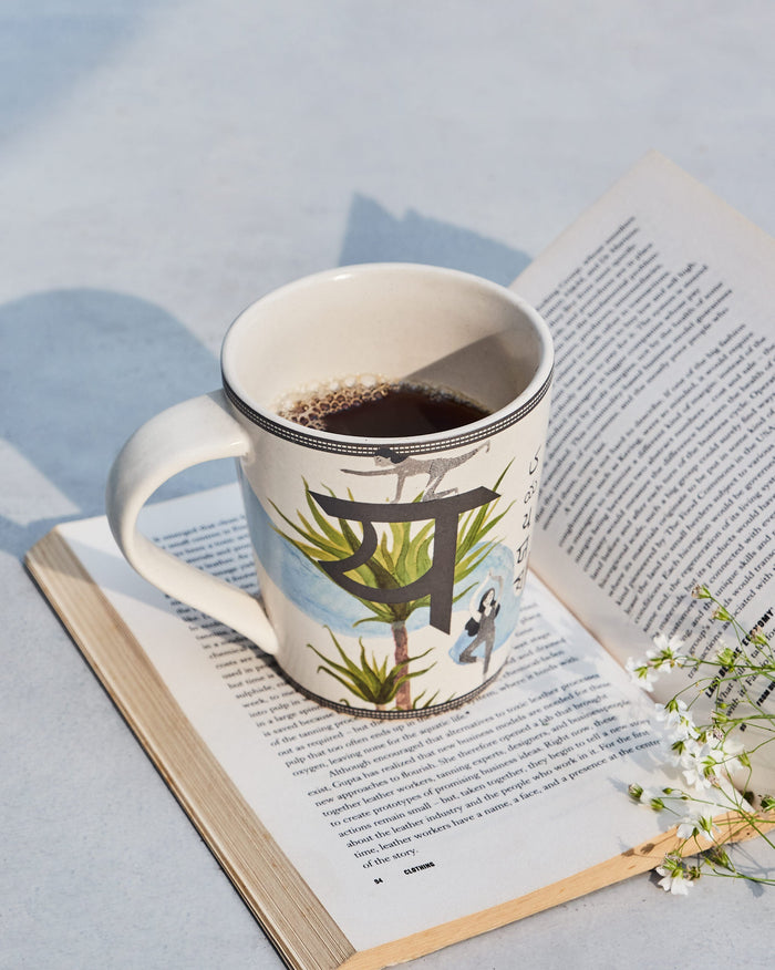 Cream Yoga alphabet mug with a Devanagari letter and yoga figure, filled with coffee.