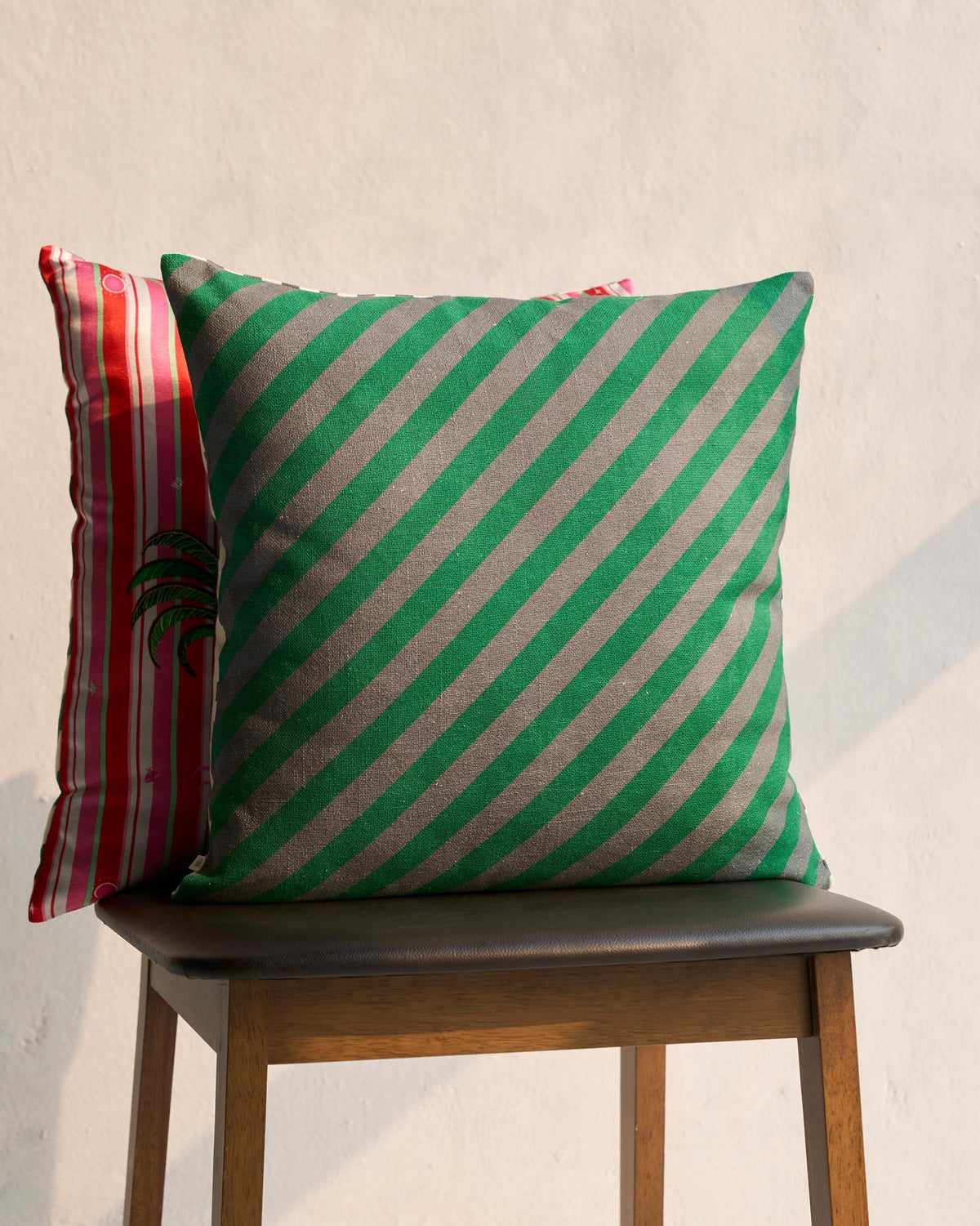 Areca Cushion Cover behind a green striped cushion on a wooden chair.