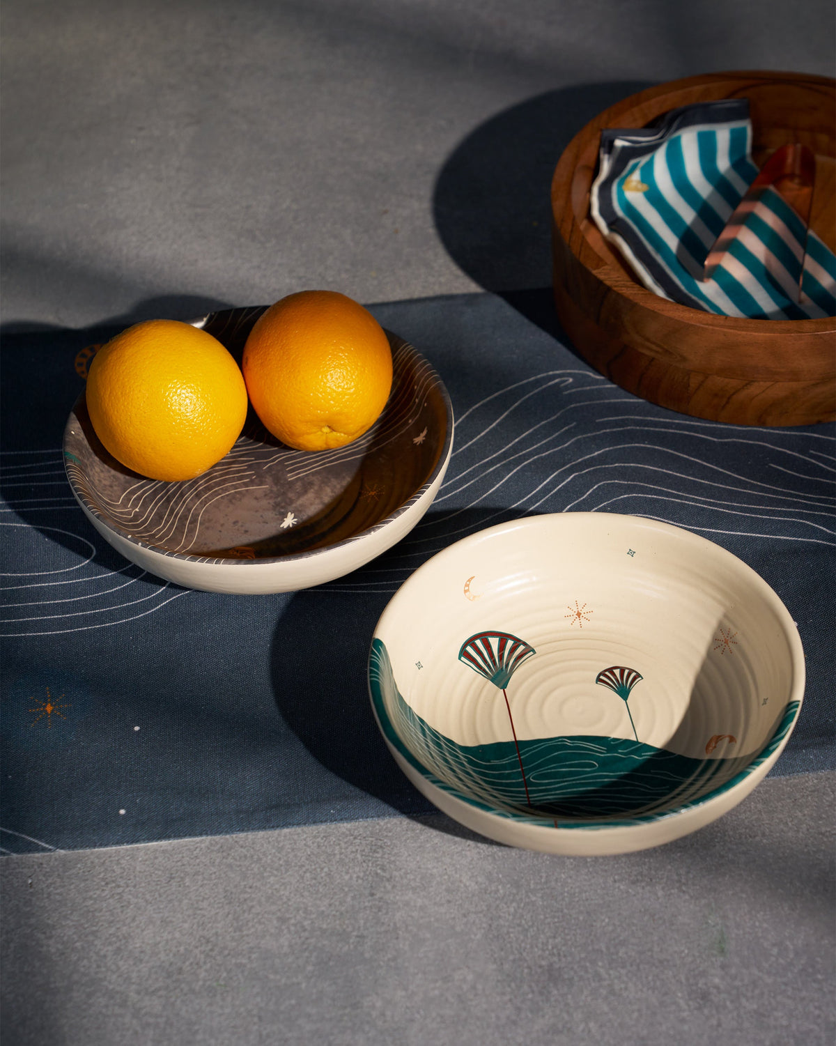 Cream Pacific shallow bowl with teal waves and fan motifs, seen at an angle beside a grey bowl.