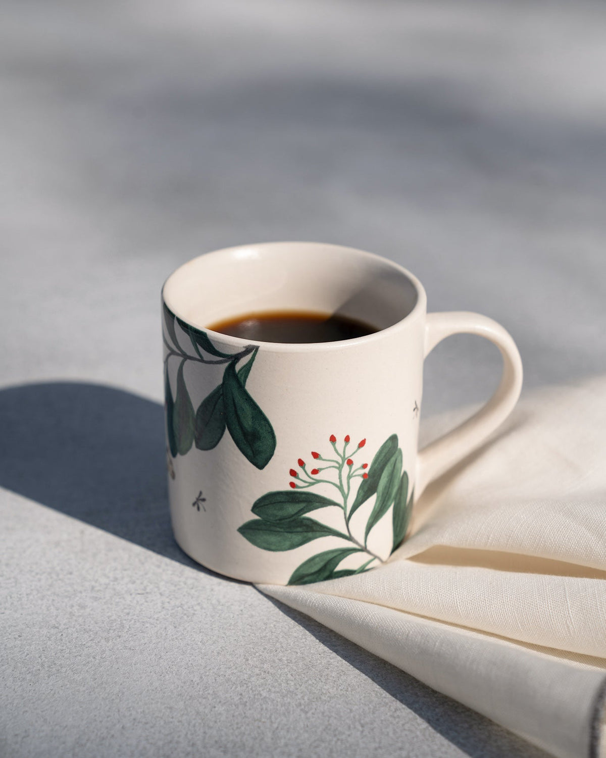 Three-quarter view of a Zuri Straight Mug with green leaves and red berries, filled with coffee.