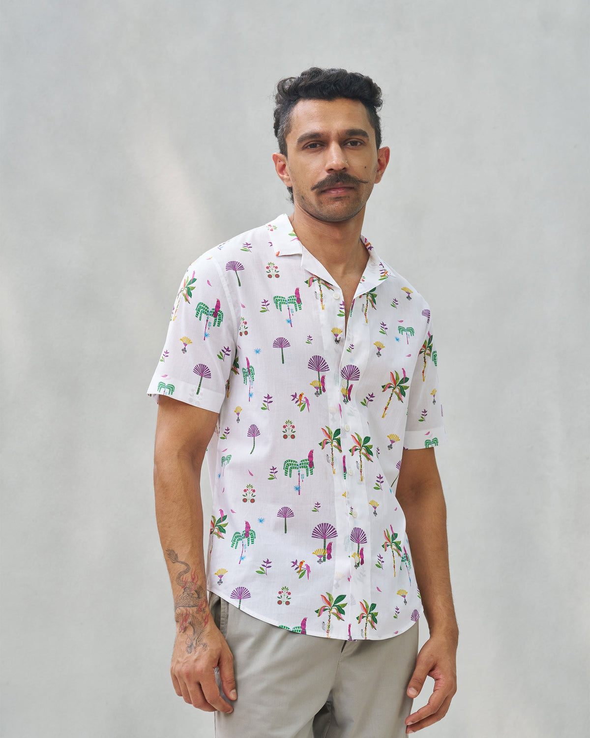 Person wearing a white Mistari shirt with multicoloured palm tree and bird motifs.