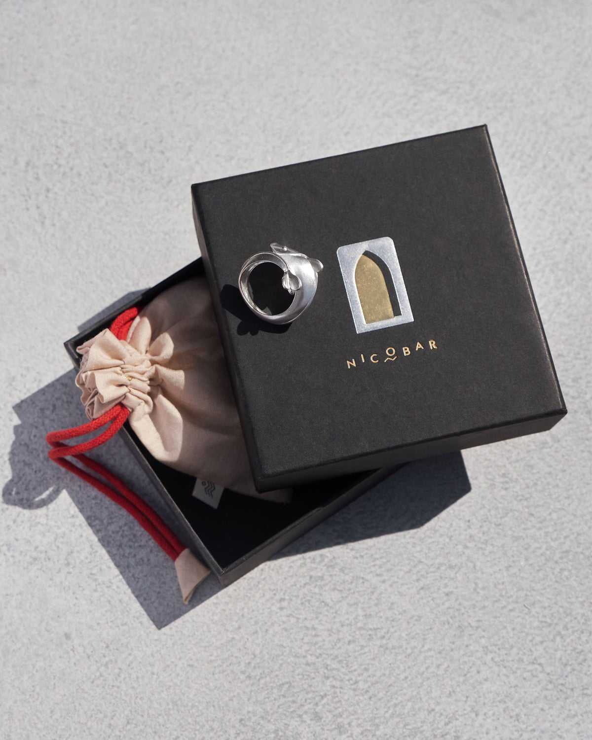 Silver Mud-Fish ring on a black Nicobar gift box beside a drawstring pouch.