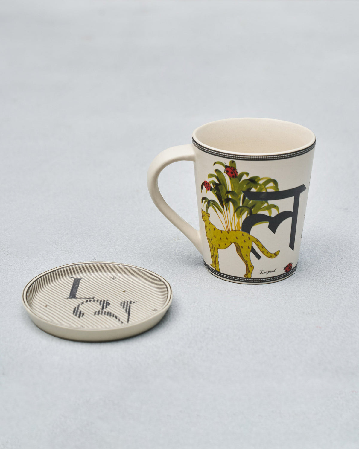 L alphabet mug with Devanagari letter and leopard motif beside coaster.