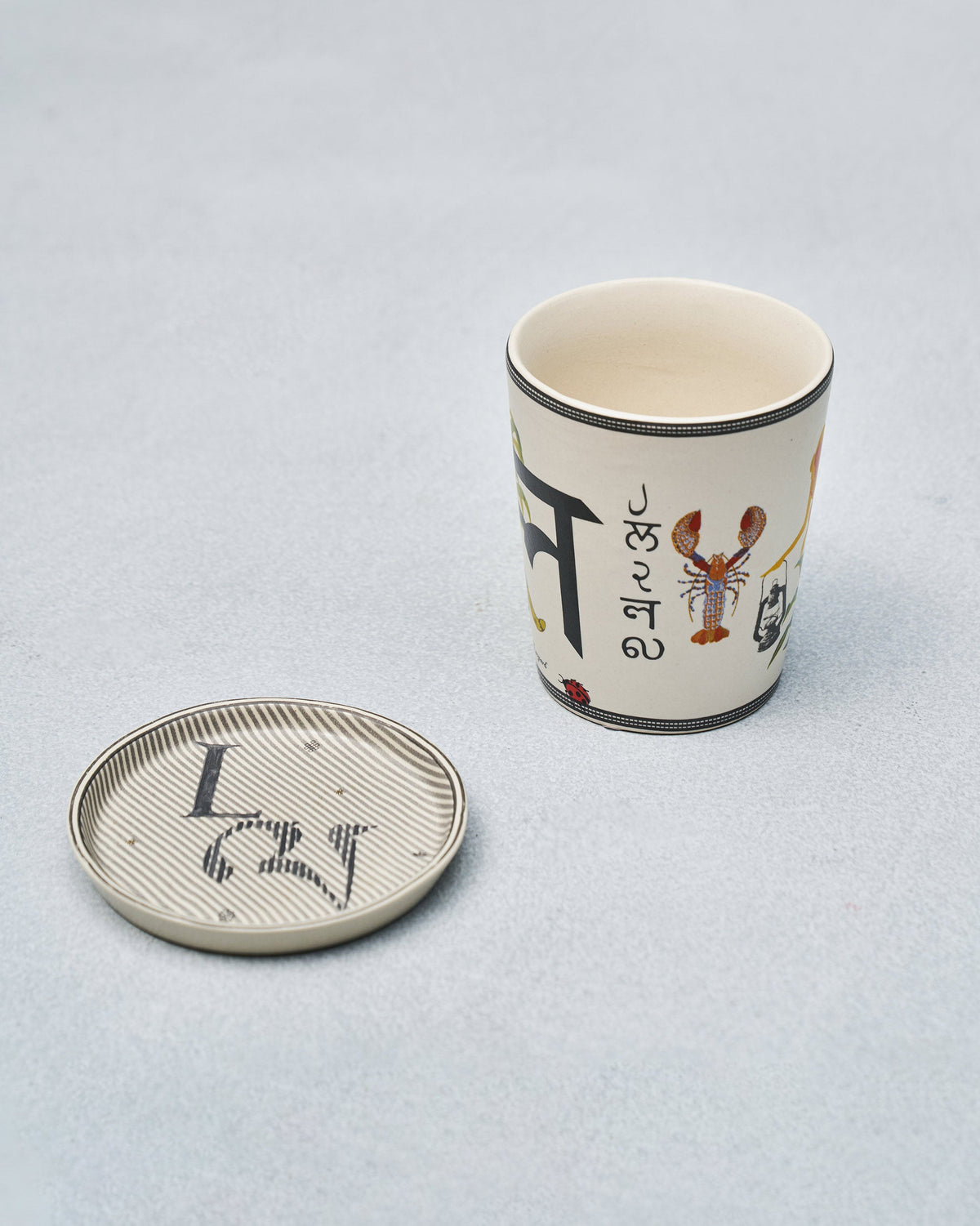 L alphabet mug with regional script and lobster motif beside coaster.