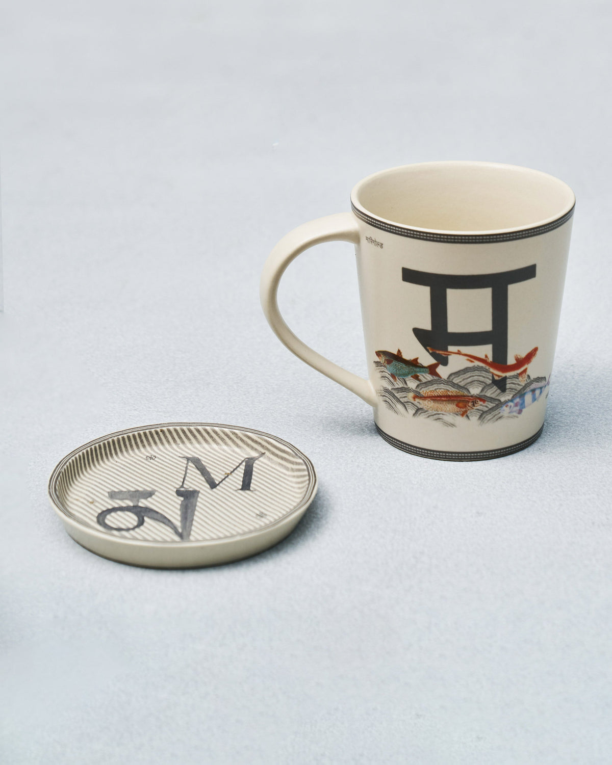 M alphabet mug with Devanagari letter and fish motif beside coaster.