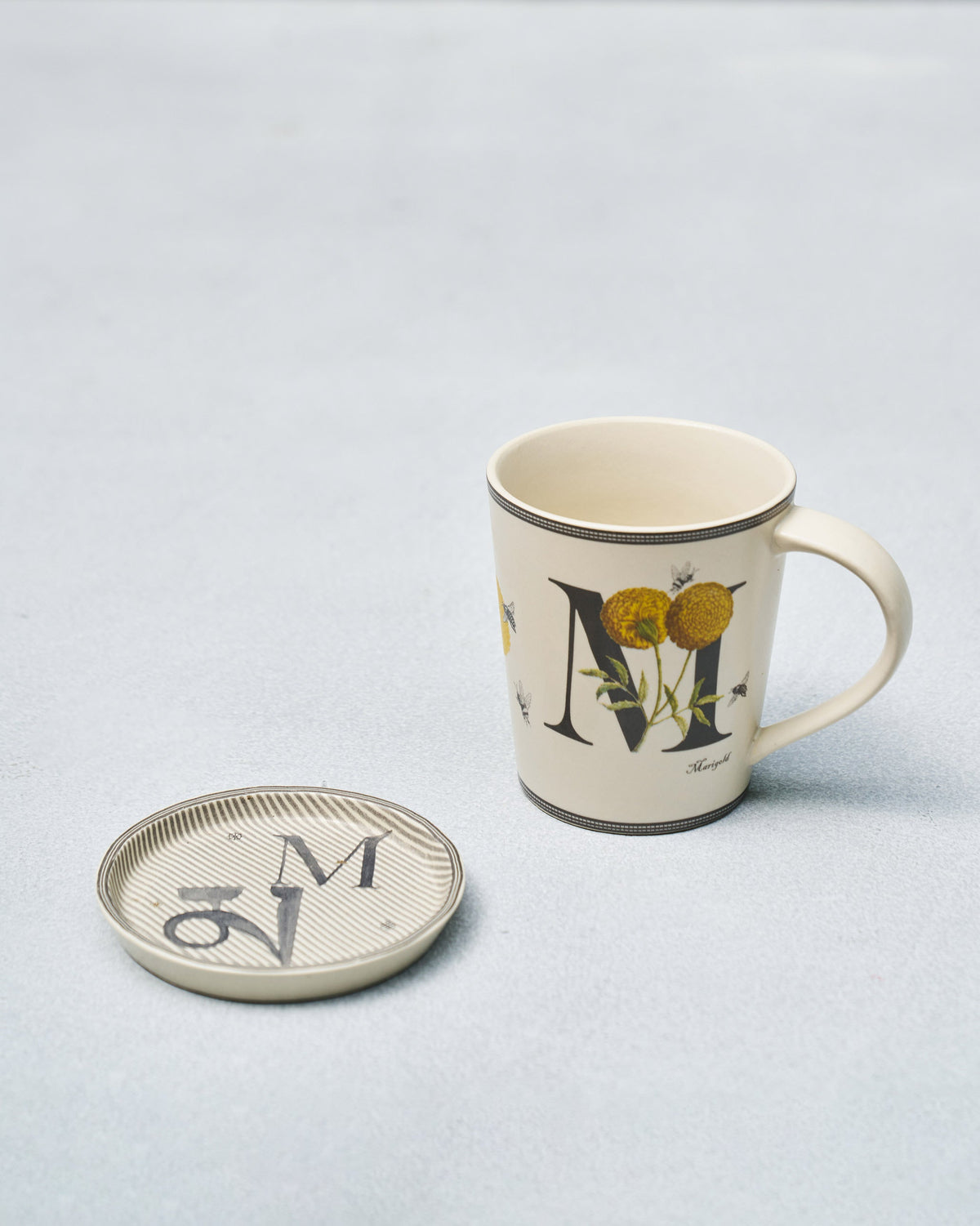 Cream M alphabet mug with marigolds and bees beside matching coaster.