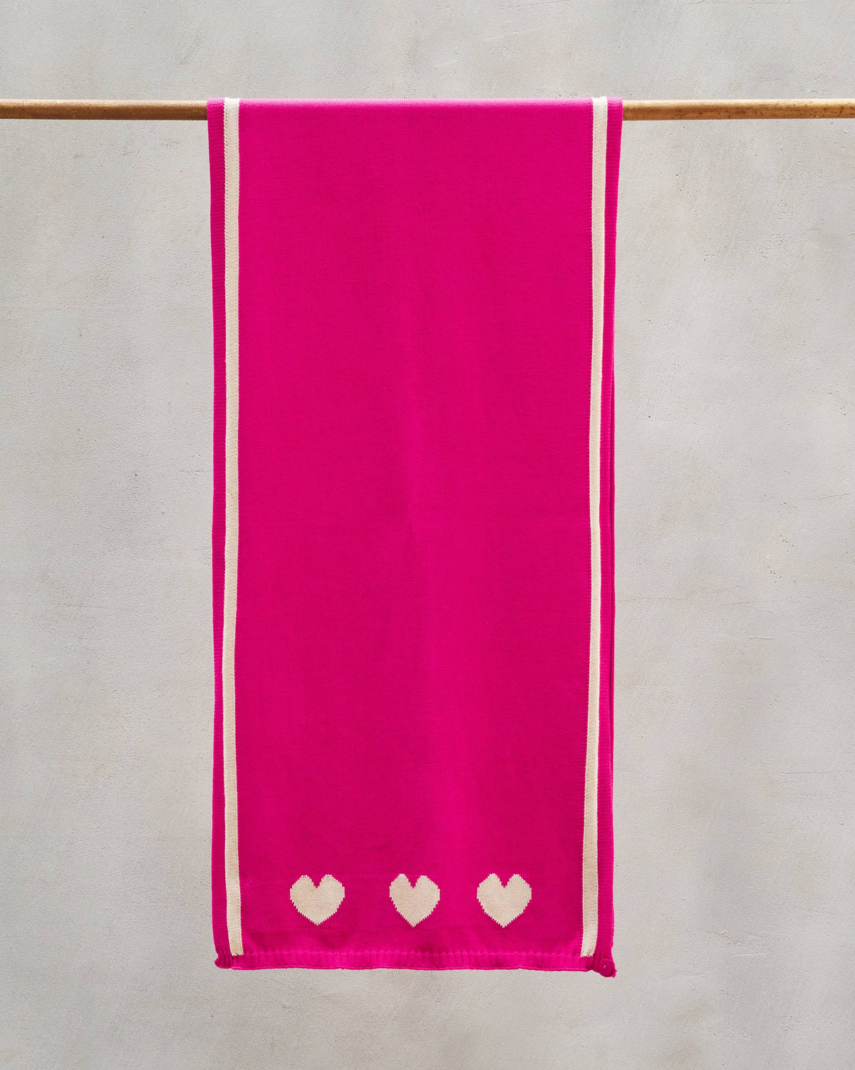 Baby Hearts Scarf in fuchsia hanging from a rail, with three cream hearts at the hem.